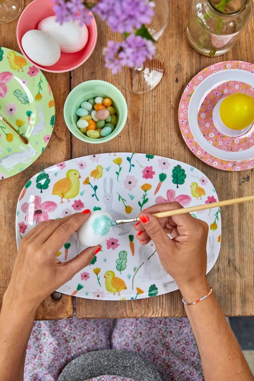Frau bemalt Ei an österlich dekoriertem Tisch mit buntem Geschirr