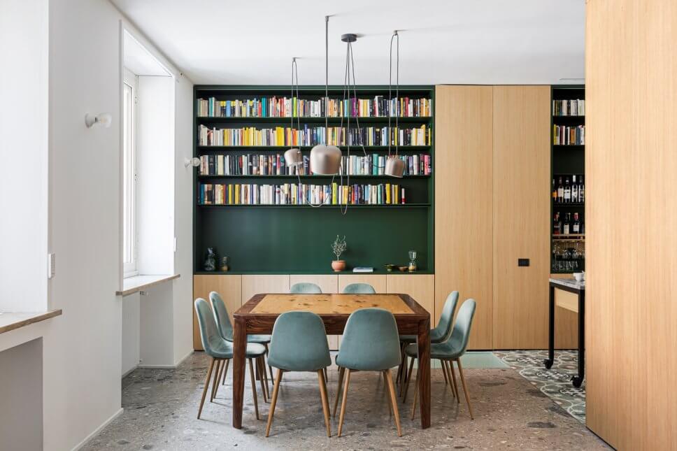 Modernes Esszimmer mit Einbauschrank aus Holz und Bücherregal auf grünem Grund mit Holztisch und acht grüngrauen Samtstühlen auf grauem Steinboden