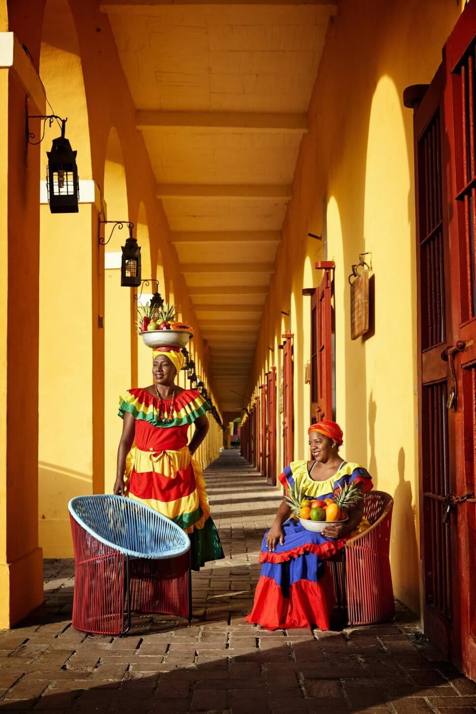 Zwei kolumbianische Frauen in farbenfrohe Tracht stehen vor zwei Sesseln aus buntem Geflecht in einem Laubengang in einer karibischen Kolonialstadt
