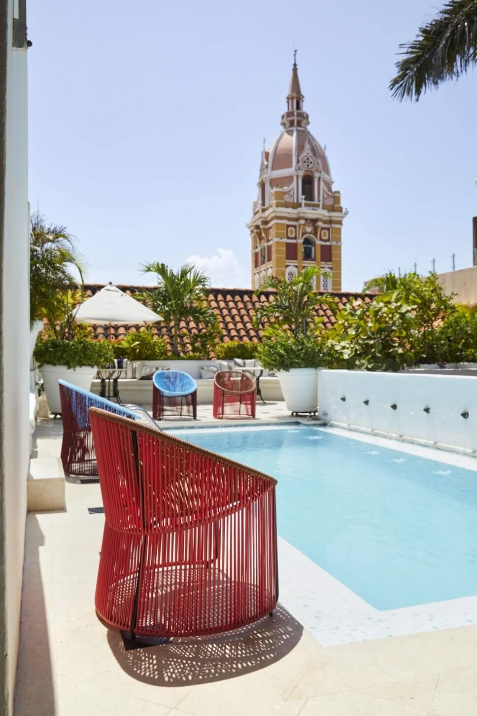 Sessel aus buntem Geflecht stehen um einem Pool in einer kolumbianischen Casa mit Blick auf Kirchturm im spanischen Kolonialstil