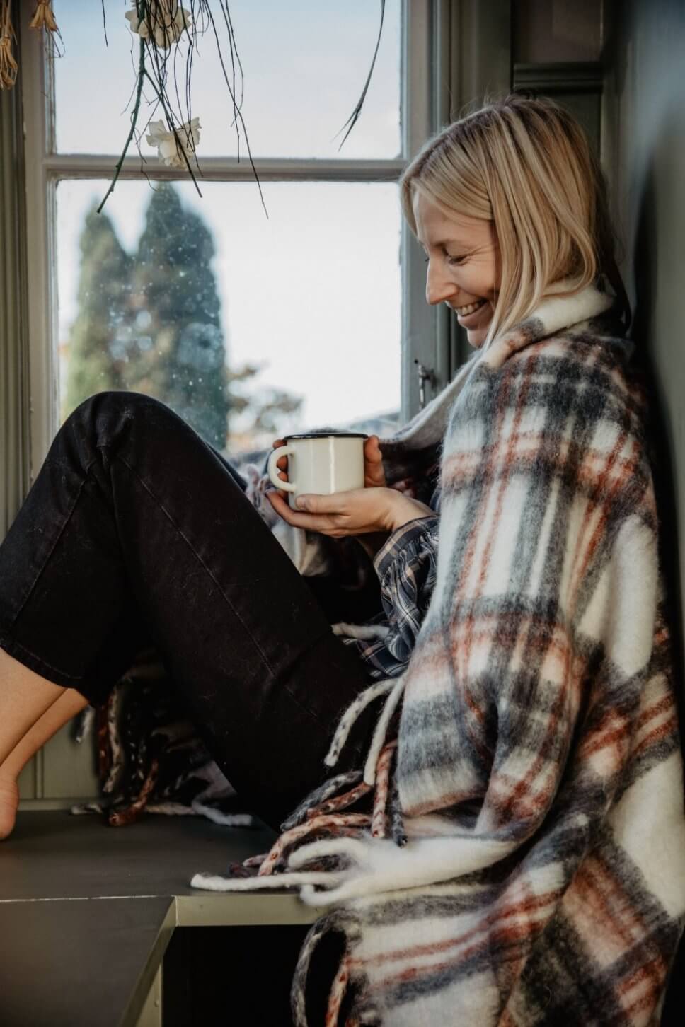 Frau sitzt mit Wolldecke und Kaffeebecher auf Fenstersims in Flur im schwedischen Landhausstil