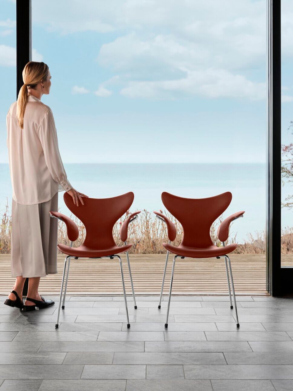 Frau blickt aus raumhohen Fenstern auf Meer, neben ihr stehen zwei modern Stühle, skandinavische Designklassiker