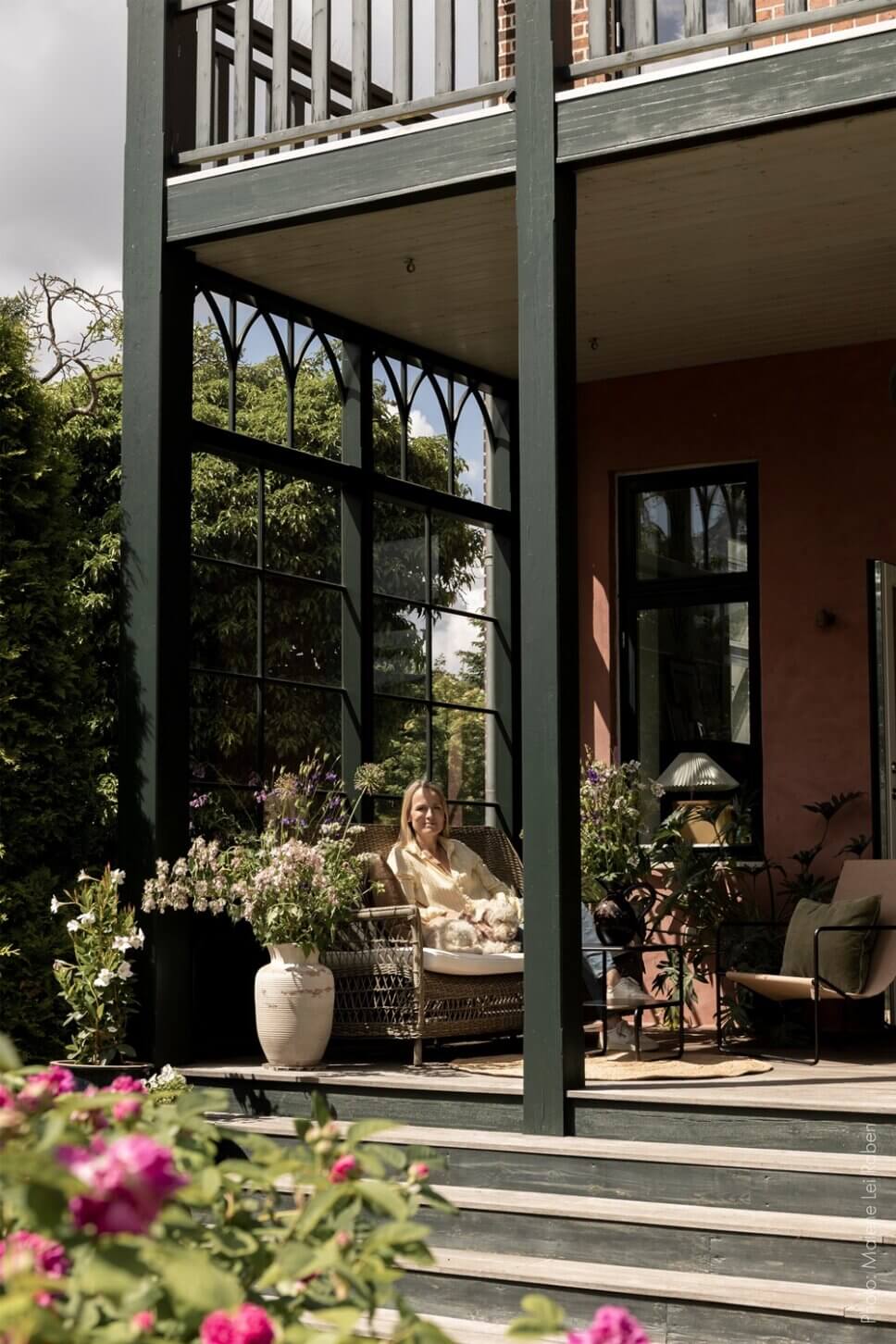 Behaglich mit Korbsofa und Esstisch eingerichtete Veranda einer alten Villa mit großem Garten. Eine Vase mit üppigem Blumenstrauß steht auf dem Tisch