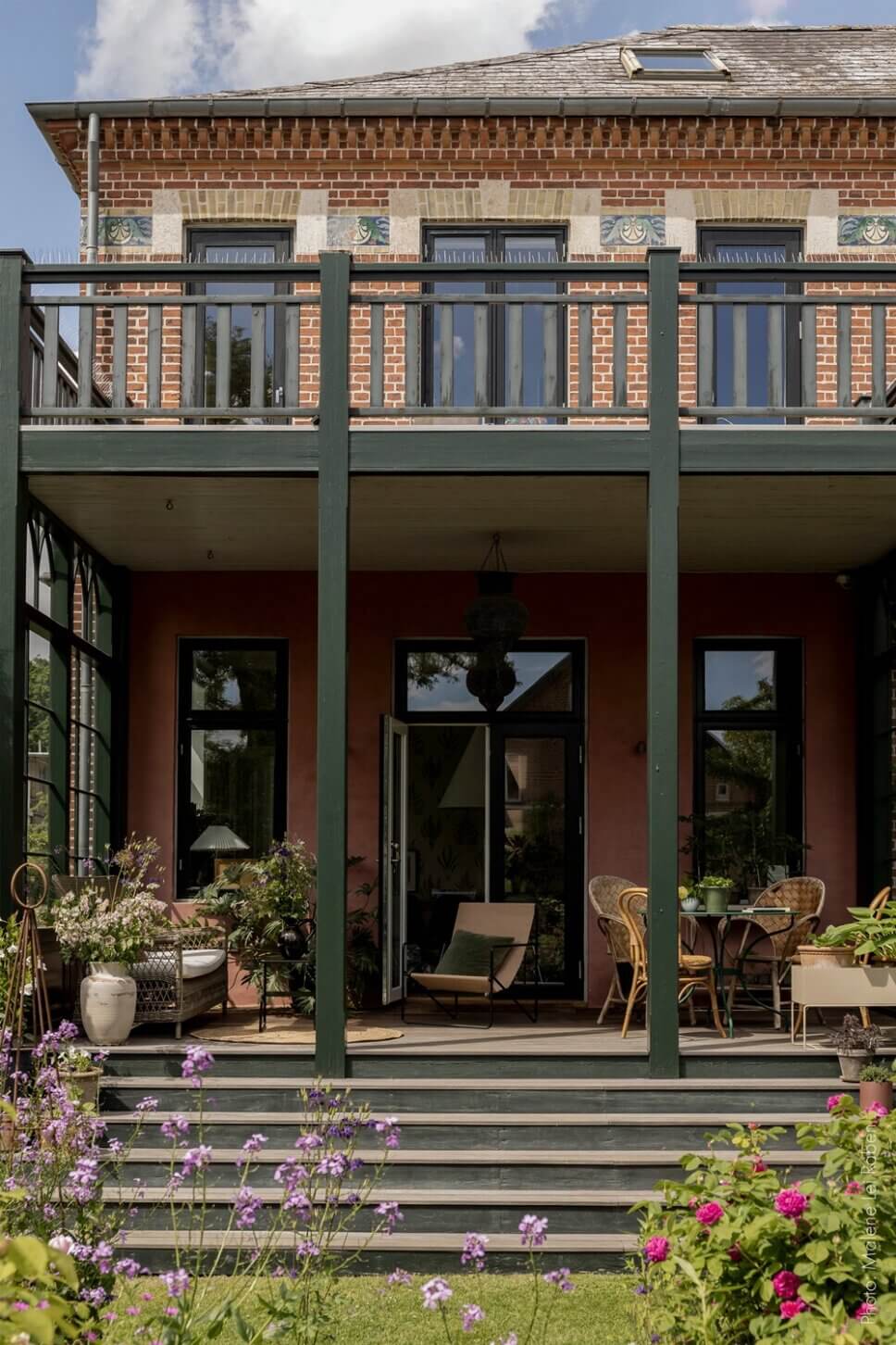 Behaglich mit Korbsofa und Esstisch eingerichtete Veranda einer alten Villa mit großem Garten. Eine Vase mit üppigem Blumenstrauß steht auf dem Tisch